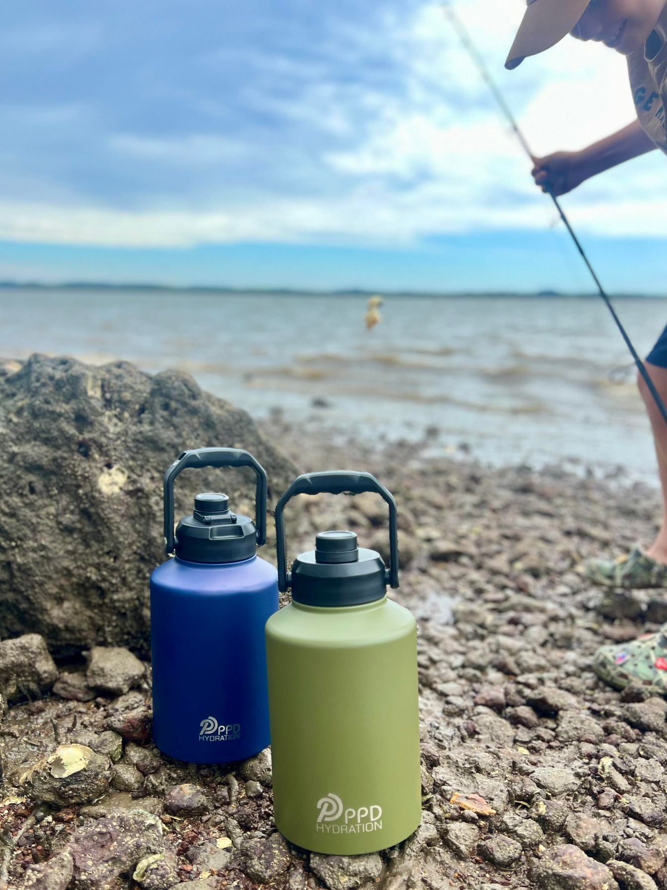 Two water bottles, one blue and one green, on a rocky surface with a person fishing in the background.