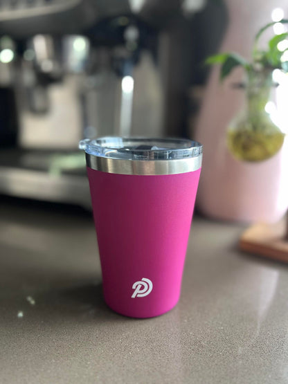 A pink stainless steel travel cup with a magnetic lid, displayed on a countertop. There is a plant in the background and a blurred object in the far right corner.