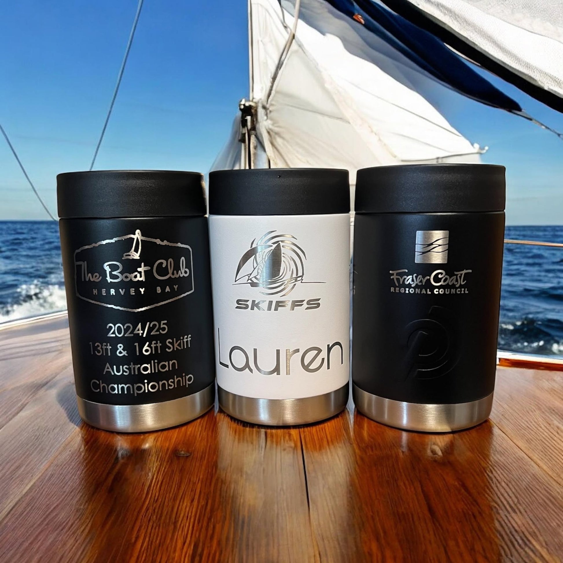 Three commemorative can coolers with personalized engravings, placed on a wooden surface with a sailboat in the background. The coolers are black, white, and green, each featuring a different name and the Skiffs logo.