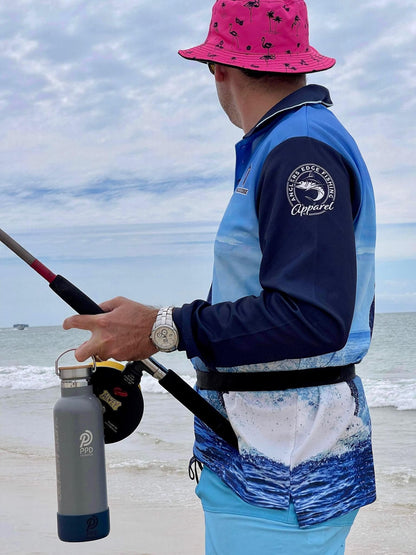 A fisherman stands by the water, holding a fishing reel and a 750 ml grey Power Drink Hydration bottle, gazing at the seawater. The sky is bright and clear.